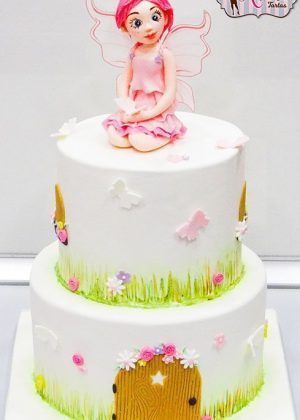 Tarta hada niña con figura de hada rosa sentada sobre una tarta blanca con puerta encantada y flores