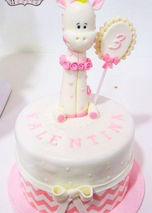 Tarta animales jirafa blanco y rosa con jirafa grande vestida de rosa sosteniendo pirulí con número 3 y nombre VALENTINA en la superficie