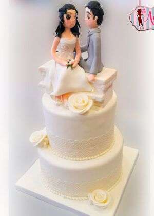 Tarta de boda con encaje y rosas de dos pisos en blanco con patrón de encaje, rosas blancas y figuras de novios sentados en un banco
