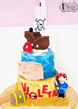 Tarta niña pirata de varios niveles con barco pirata, bandera de calavera, mapa del tesoro, loro rojo y figura de niña pirata sobre base de arena