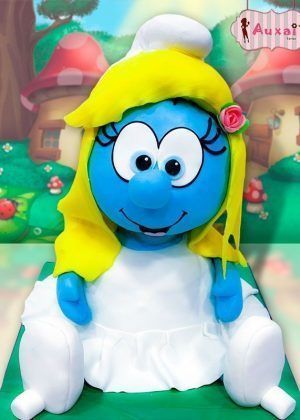 Tarta 3D Pitufina figura completa azul con cabello rubio, gorro blanco y vestido blanco sentada sobre base verde