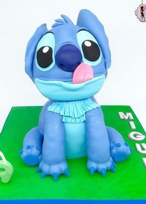 Tarta Stitch 3D con figura esculpida de Stitch sentado, lengua rosa fuera, ojos grandes y base verde con nombre MIGUEL en letras blancas y vela número 9
