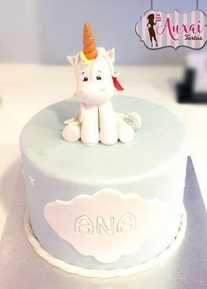 Tarta unicornio azul cielo con figura 3D de unicornio blanco sentado, cuerno dorado y nube frontal con nombre ANA en letras en relieve