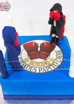 Tarta ring de boxeo: estructura multi-nivel con lona azul, cuerdas blancas, guantes de fondant rojo y azul, campana dorada y postes de soporte detallados
