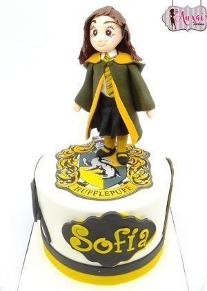 Tarta Harry Potter Chica personalizada con fondant amarillo dorado y detalles en negro. Escudo de Hogwarts modelado en fondant. Formato individual con nombre personalizado del celebrante. Acabados profesionales de categoría mundial.